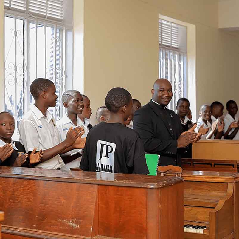 A group of male students and a Jesuit applaud in front of a room with donated pianos.