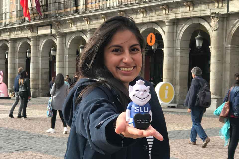 Female Madrid student holding hand out with squishy Billiken, with arches in background