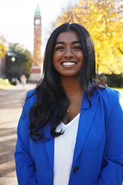 A headshot photo of Reueline Arulanandam with a clocktower and trees in the background.