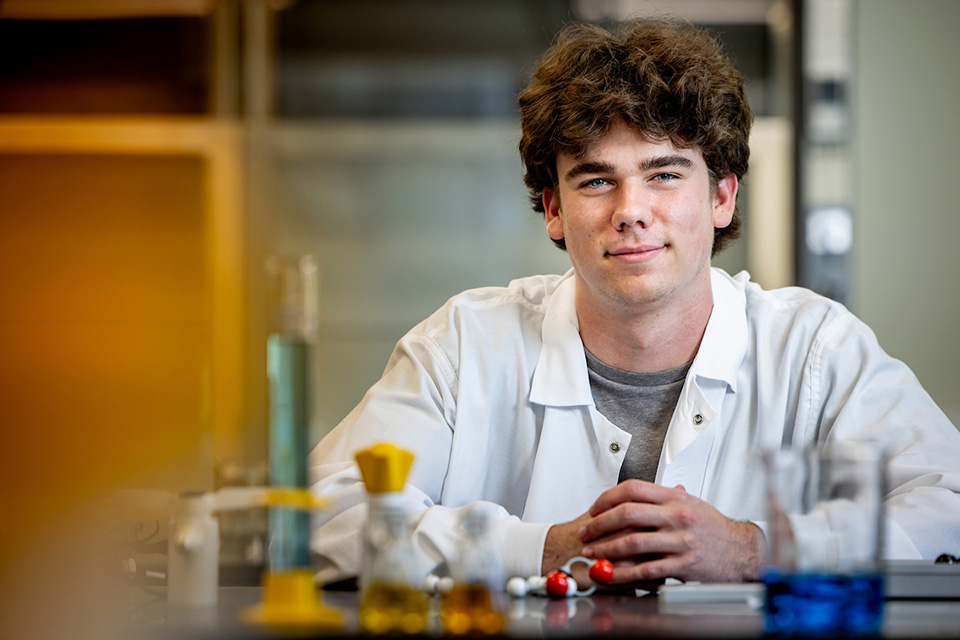 A male student poses for a photo wearing a white lab coat sitting in a science lab.