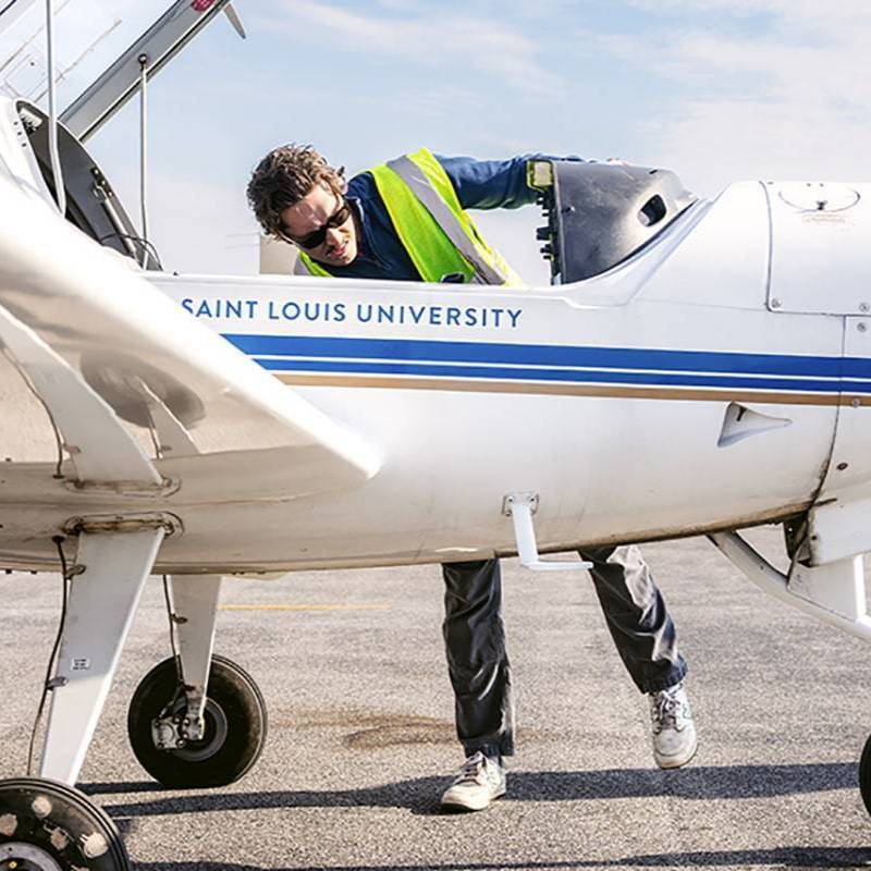 A student wearing a reflective safety examines the cockpit of an airplane that says Saint Louis University on the side. 