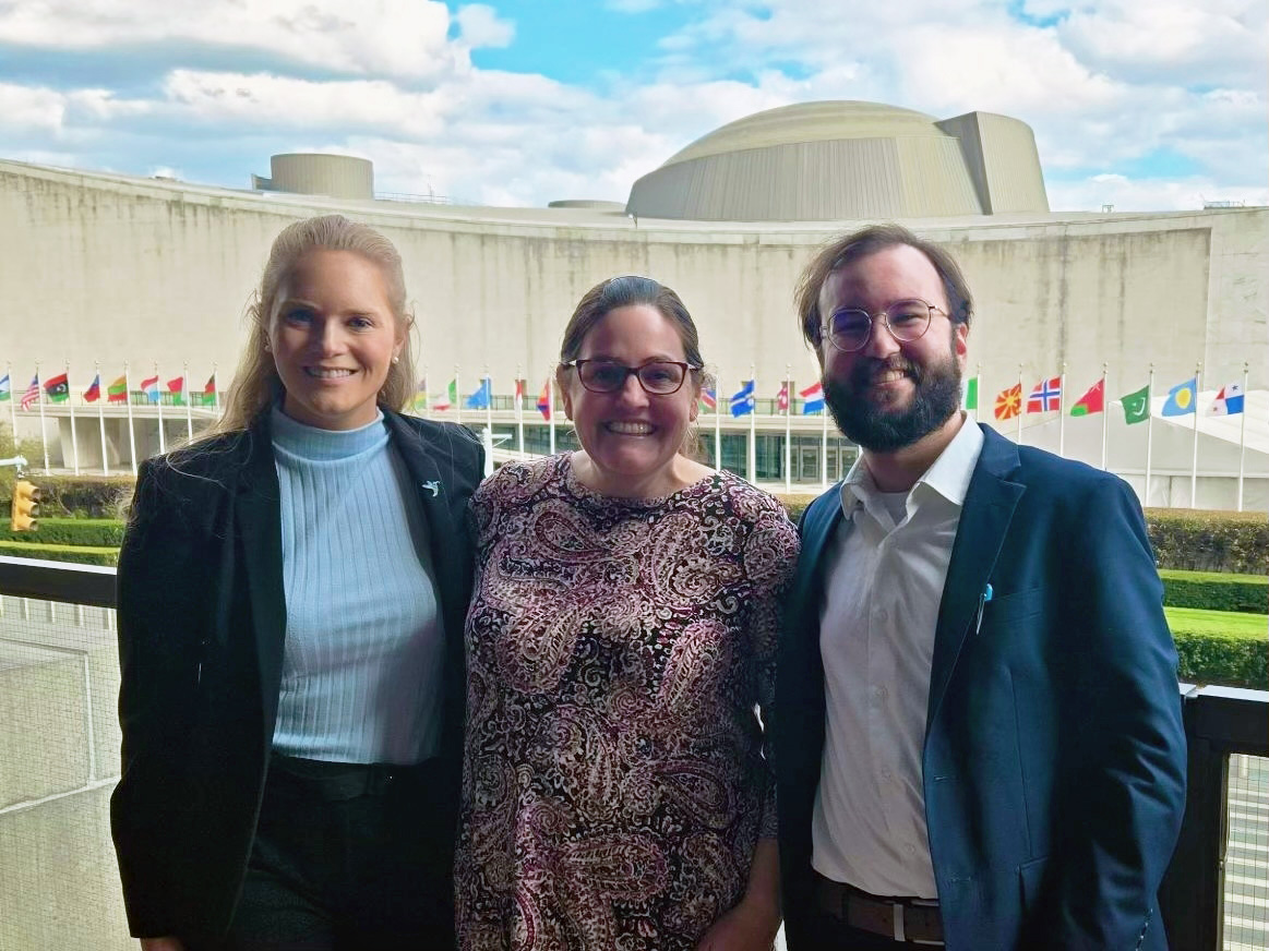 Human Rights at Home Clinic students with Professor Lauren Bartlett pose in front of the United Nations building in New York, NY clinic students and professor in front of a building lined with flags.
