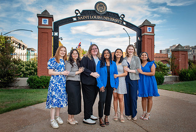 Seven professionally-dressed young women standing in front of the SLU archway