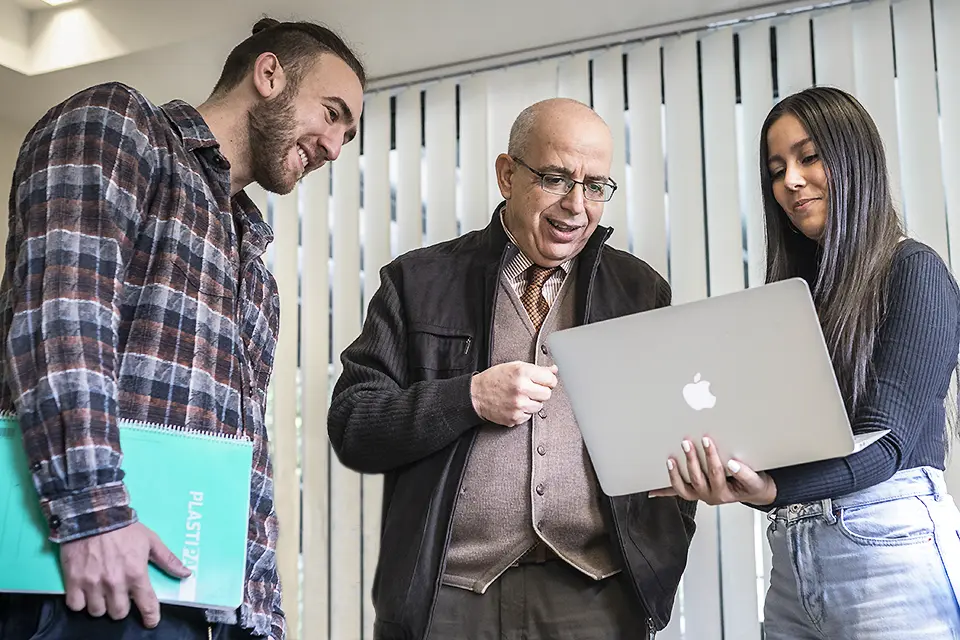 Two students talk to a professor while looking at a laptop.