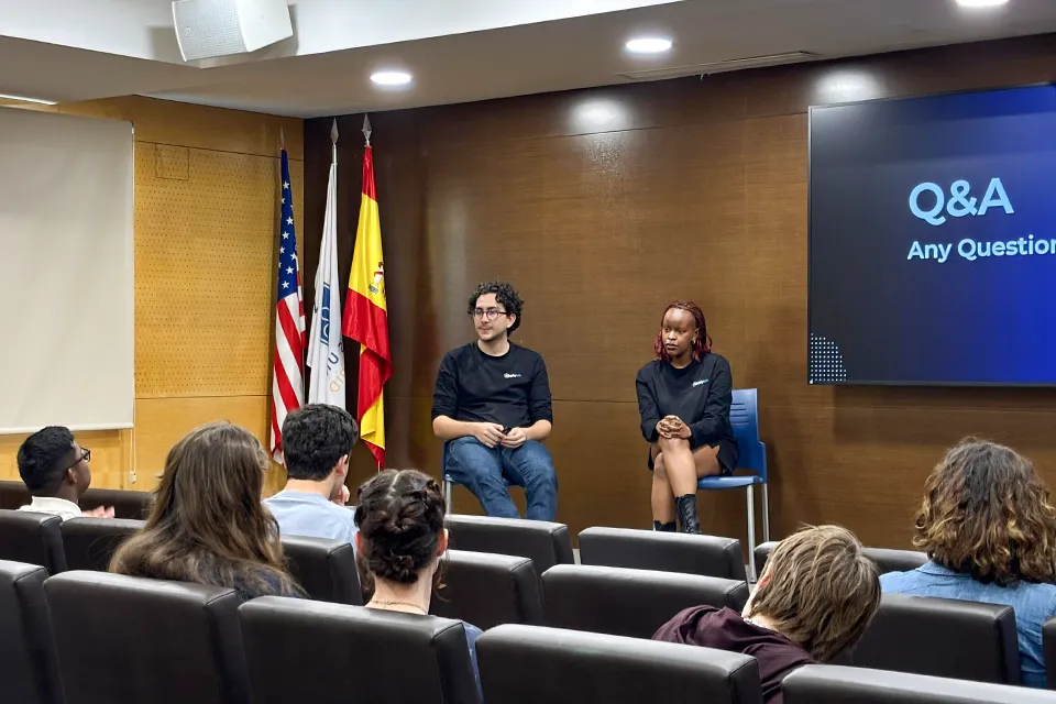 Guest speaker and a panelist sit at the front of the auditorium facing students seated in the audience with U.S., Spanish and university flags visible behind them and a presentation slide reading "Q&A – Any Questions."