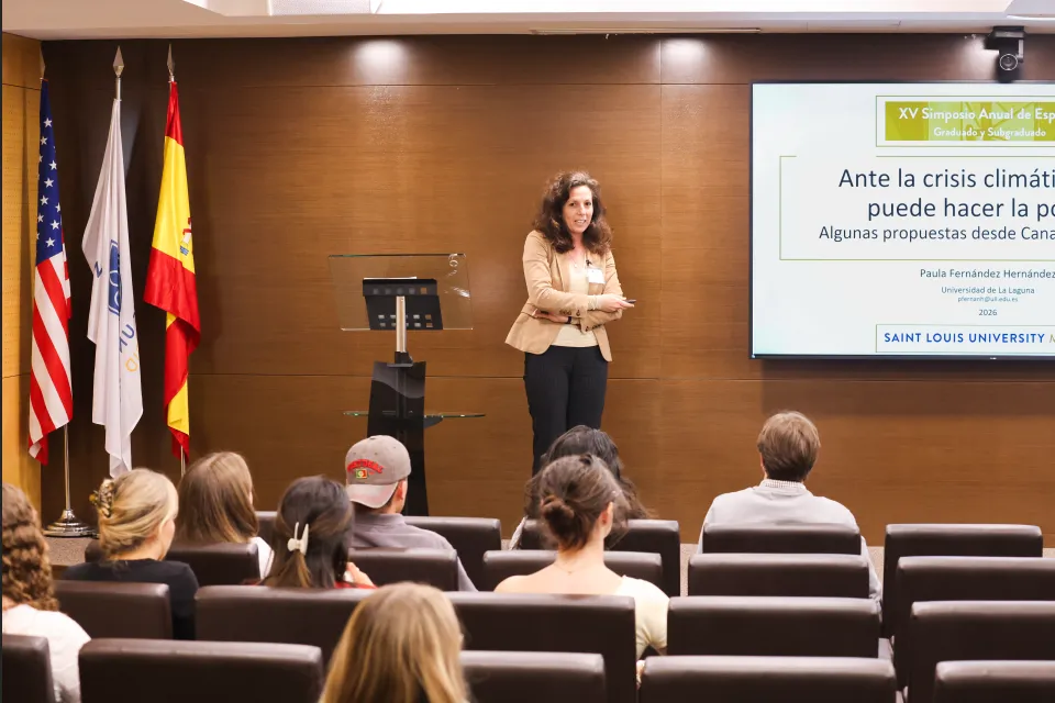 A woman speaking at the front of a lecture room, engaging a small group of students during the XV Annual Spanish Symposium at SLU-Madrid.