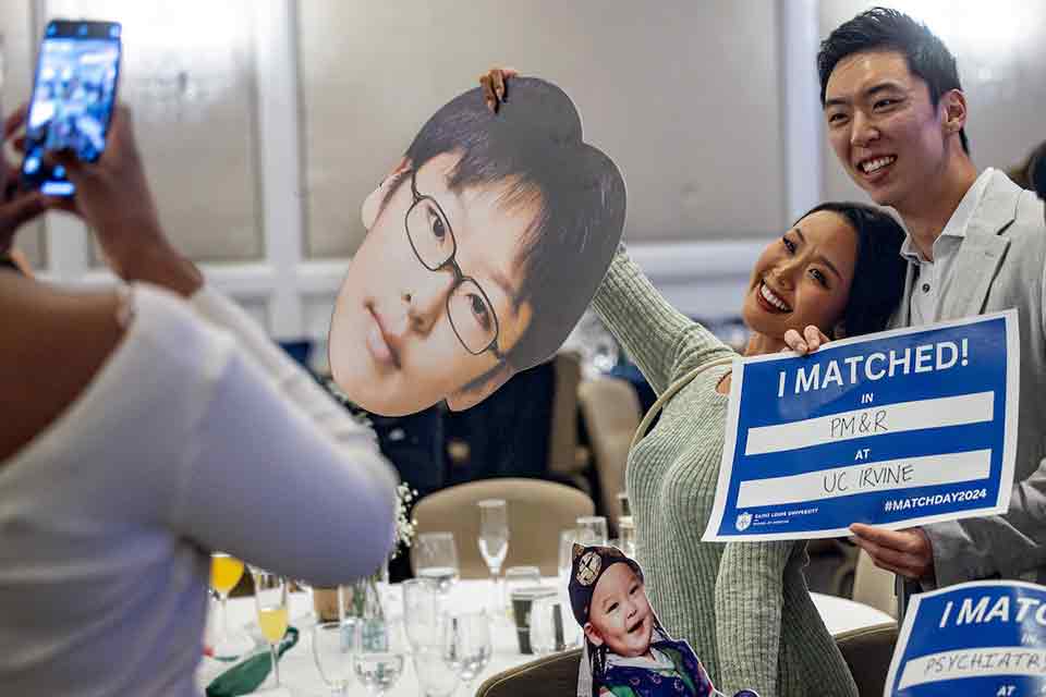 A student holds a sign that reads "I matched in PM & R at UC Irvine." A family member hugs him while holding a cardboard cutout of his head.