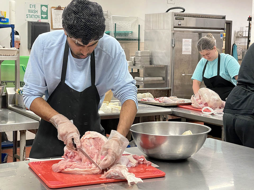 A man cuts meat on a red cutting board next to a stainless steel bowl on a stainless steel countertop. A woman prepares a turkey in the background.