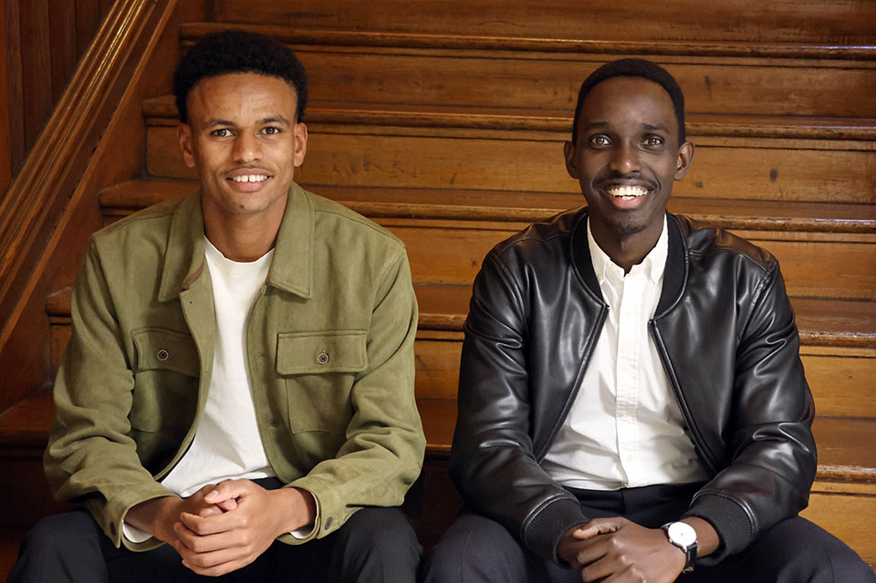 Two men pose for a photo. They sit on wood stairs. 