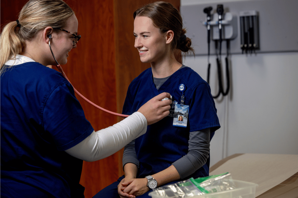 Two nursing students in navy scrubs in the lab. One of the students is using a stethoscope to listen to the other student's heart.