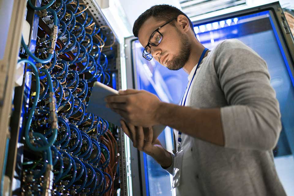 Cybersecurity professional view data on a tablet near a wall of wires