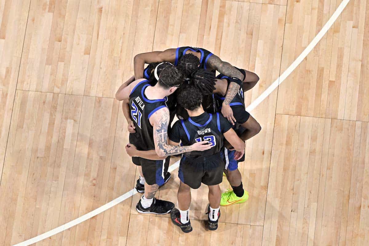 Members of the men's basketball team stand in a huddle in the middle of the court during a game.