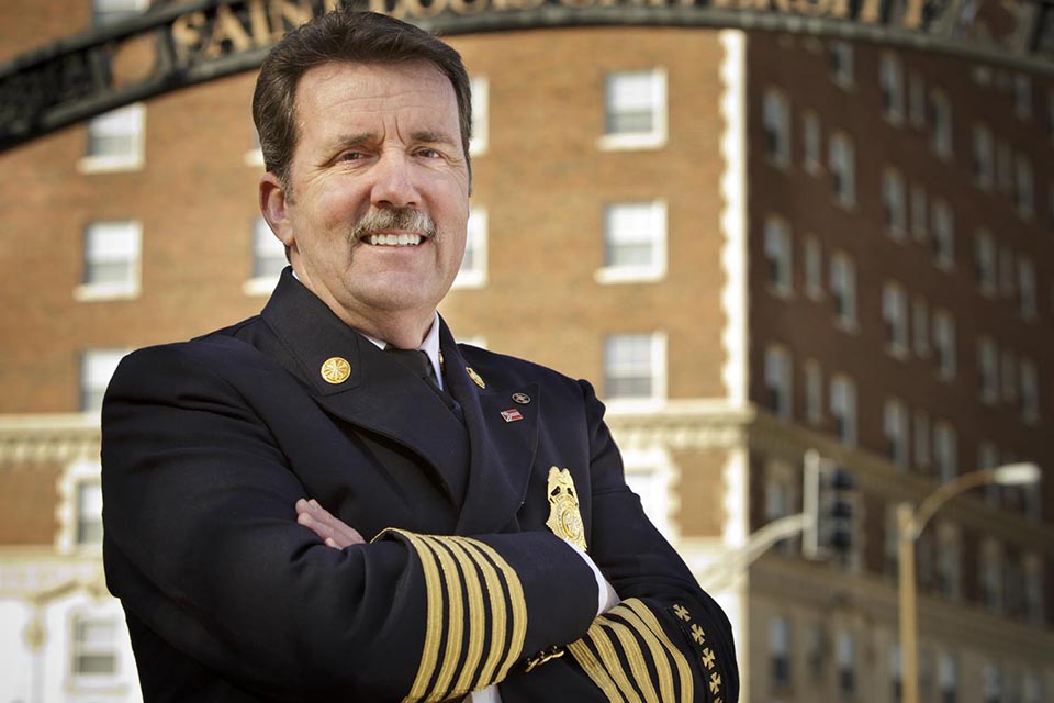 A man in a fire chief's uniform stands with arms folded in front of the archway reading Saint Louis University.