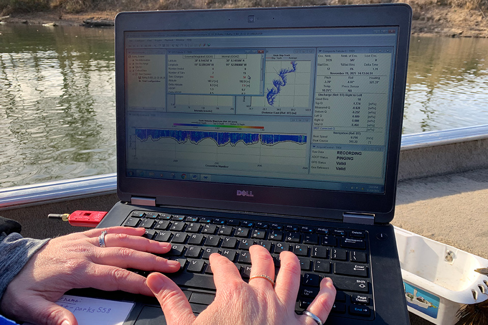 A pair of hands type on a laptop keyboard, while the screen shows charts, graphs and data sets, situated with a river in the background.