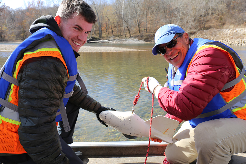 Two men wearing life jackets and reflective gear sit in a small boat on a river. They are holding a small piece of equipment on a rope. Rocky shore line with bare trees is visible in the background.