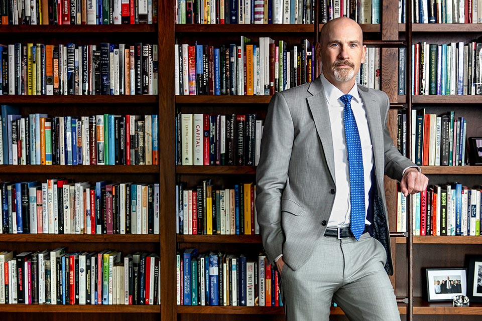 A bald white man in a grey suit stands against a wall of books